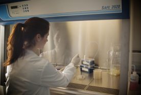 researcher in white coat working with pipette under a protective hood