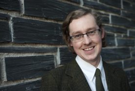 John Tredinnick-Rowe smiling to the camera leaning against a wall