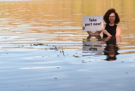 PhD researcher Elitsa Penkova stands waist deep in a calm sunny river holding a sign saying 'take part now!'