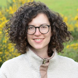 Elitsa has brown curly hair and wears glasses as she smiles for the camrea outside with yellow gorse flowers behind