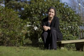 Lora sits on a bench outside in the sun against a green hedge smiling