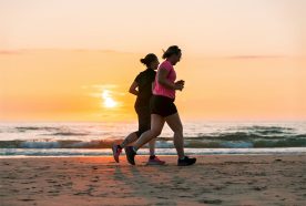 Two women run along flat sand close to the water on a calm beach as the sun sets behind them.