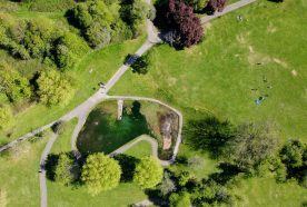 A green park seen from above with small paths winding through it and a pond to the left centre of the image.