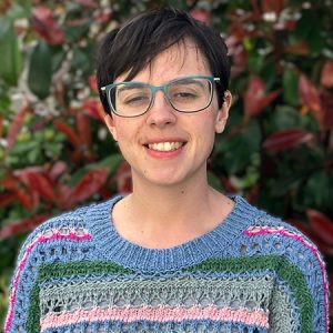 Charlotte wears a blue and green knitted sweater standing in front of a green bush with red flowers.