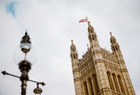 An angled view of the spire of Westminster Abbey set against a bright but cloudy sky, an old lamp post sits in frame to the left.