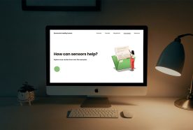 An Apple iMac computer on a dimly lit desk with the Sensors for Health Homes website loaded. A lamp and pot plant sit either side of the screen.