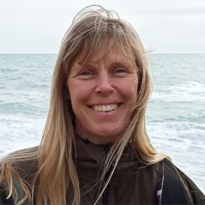 Gemma smiles for the camera with head and shoulders in view and the ocean in the background.