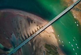 A bridge crossing between two sandy outcrops and over a green river is viewed from very high above.