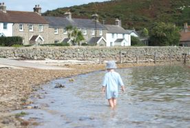 A toddler in a blue one piece walks in shallow water in a calm harbour with sea wall and cottages in the background.