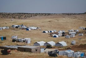 A collection of temporary tents and shelters set in a brown yet grassy undulating landsape.