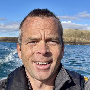 Charlie smiles as he takes a close-up selfie of his face from a boat with blue sea, sky and cliffs in the background