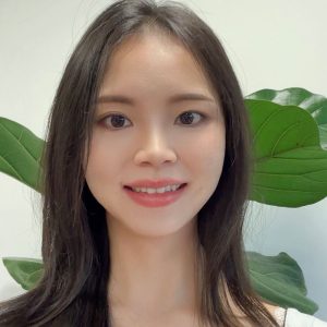 Siyu smiles for the camera in front of a white wall with green plants.