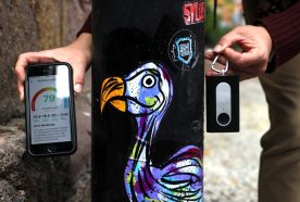 A close up of a lamp post with a colourful dodo painted on - on either side two hands hold a small silver sensor and phone with air quality data display
