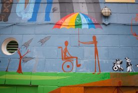 A mural painted on a blue wall shows a rainbow coloured umbrella held by a standing person to shelter a person in a wheelchair.