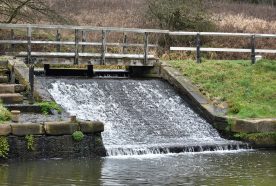A small weir separates two levels of a canal, with water hently flowing down a short slope.