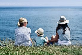 A family of three sit on a grassy cliff, with their backs to the camera, looking out on a calm blue ocean with clear skies above.