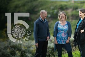 Mike, Emma, and Lora face each other, laughing, in a sunny green landscape with the 15yr logo overlain to left of image.