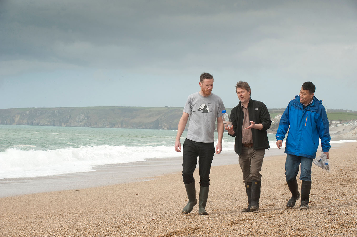 Three researchers walk along a steep sandy beach near the shoreline talking under cloudy skies.