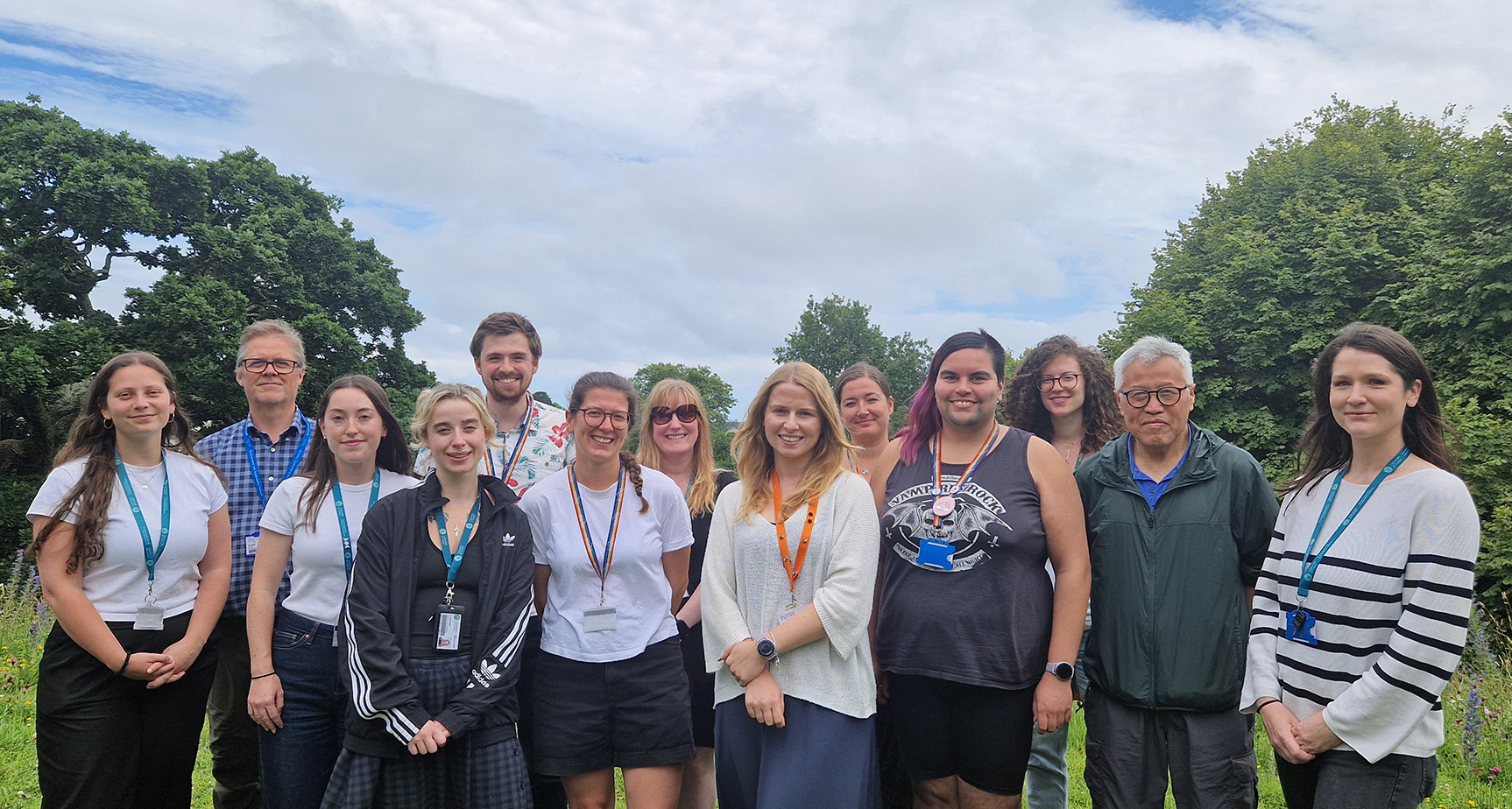 13 AMR team members assembled in a line, outside on a cloudy summers day with green trees behind.