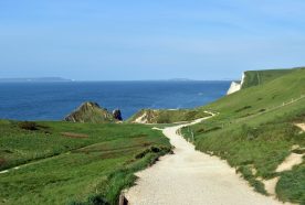 A wide gravel path snakes along a green cliff top with blue seas and skies in the background.