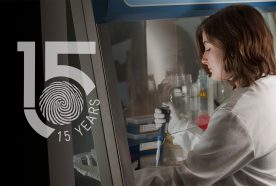 Aimme Murray sits with her arms in an enclosed glass cabinet fills vials with a pipette. The 15 year logo sits to her left.