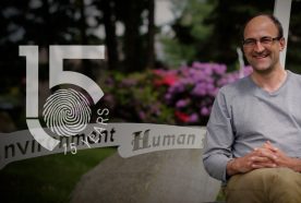 Ben Wheeler sits on a bench in summer in a grey long sleeved t-shirt with purple flowers behind. The 15yr logo is overlaid to the left of the image.