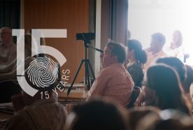 People at a conference look towards a speaker (not in view) as bright light bleads through a window behind. The 15 yr logo sits to the left of the image.
