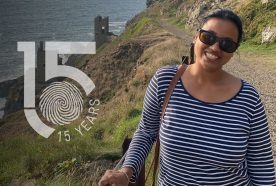 Monika Walia stands on a gravel path above a high cliff with rough blue seas and the ruins of a tin mine behind. The 15yr logo is left of the image.