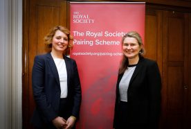 Ann Conzatti and Jane Kirkham MP smile next to a red banner in a wood panelled room.