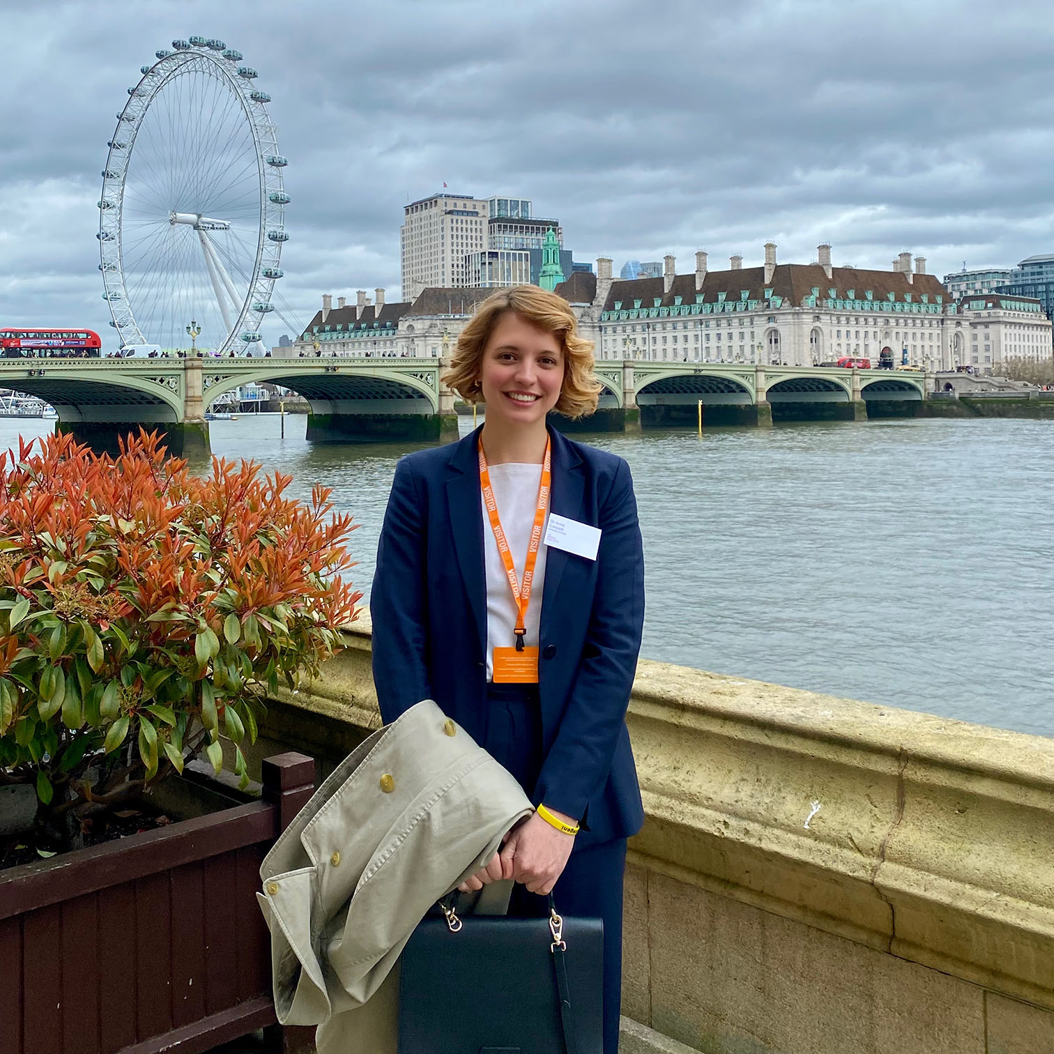 Anna Conzatti wears a smart suit and stands on the bank of the Thames with the London Eye in the background.