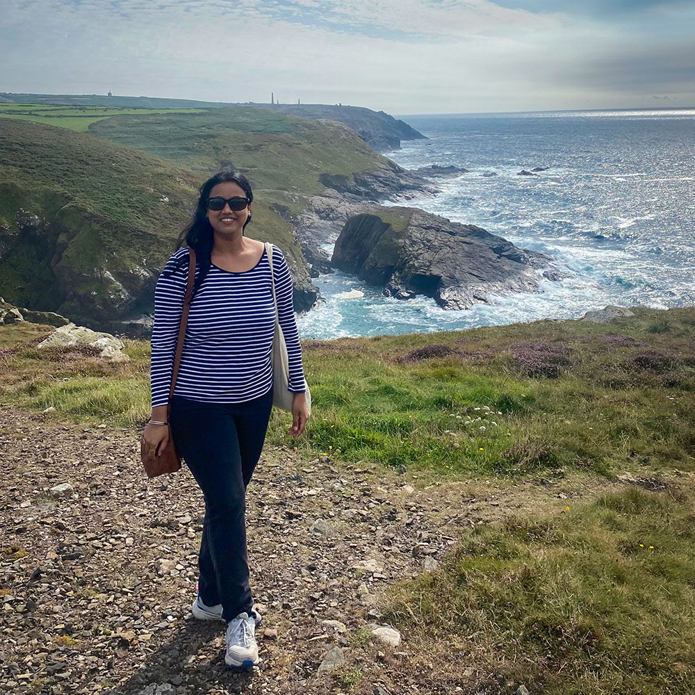 Monika Walia stands on a gravel path above a high cliff with rough blue seas below.