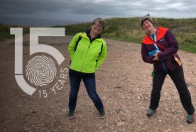 Two of the Centre's researchers, wearing coats on a grey day, stand in a clifftop car park, 2m apart, leaning towards each other.