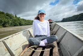 Anel sits at the front of a long narrow boat as it rides along a wide brown river flanked by rainforest.
