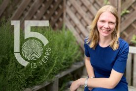 Gesche smiles of the camera sitting outside on a wooden bench with green plants to the left. The 15yr logo sits to the left of image too.