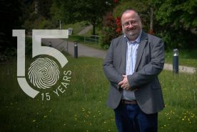 Tim taylor stands outside in a grey suit jacket with arms crossed smiling. Green grass and shrubs are behind him with the 15yr logo to left of the image.